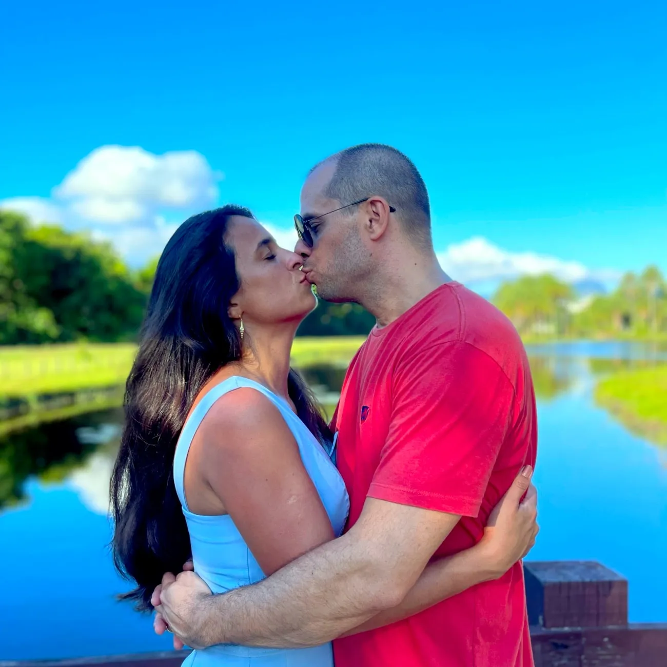 Michelle e Adriano se beijando à beira de um lago, sob o céu azul