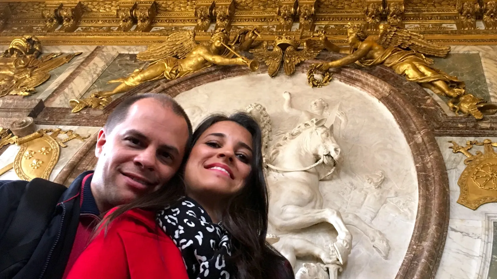 Michelle e Adriano numa selfie, sorrindo, num salão de paredes douradas com esculturas
